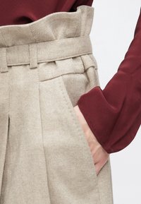 Pantalon beige taille haute avec plis et détail ceinture, associé à un top à manches longues rouge foncé, main dans la poche.