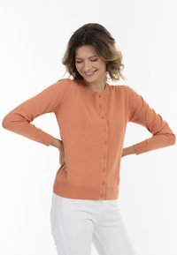 Kortärmad orange kofta med knäppning framtill, ribbat nederkant och ärmslut, tillverkad av ett mjukt material. Bärs över vita byxor.