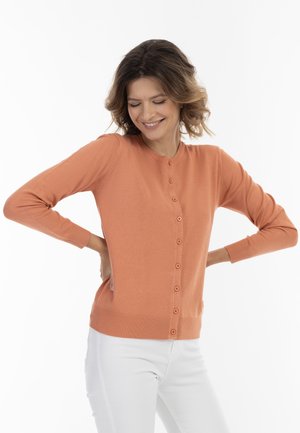 Cardigan scurt cu mâneci, de culoare portocalie, cu închidere cu nasturi, margine și manșete cu o textură ribbed, realizat dintr-un material moale. Purtați peste pantaloni albi.
