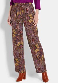 Frau in gemusterten, weiten Hosen mit Paisley- und Blumenmotiven in Rot, Lila und Senfgelb, kombiniert mit rostfarbigen, hohen Stiefeln.
