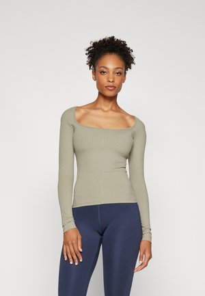 Long-sleeve top in olive green ribbed fabric with a square neckline and fitted silhouette, paired with navy blue leggings.