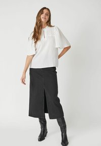 Top bianco a maniche corte con dettaglio plissettato e scollatura a chiave; abbinato a una lunga gonna di jeans nera con spacco frontale, completato da stivali neri.