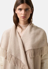 Beige vlněný šál s třásněmi, měkkou strukturou a obdélníkovým tvarem, přehozený přes ramena, vrstvený s žebrovaným svetrem.