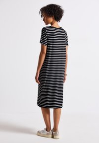 Mujer con cabello rizado, vestida con un vestido midi a rayas horizontales en blanco y negro y zapatillas blancas, de pie de lado frente a un fondo liso.