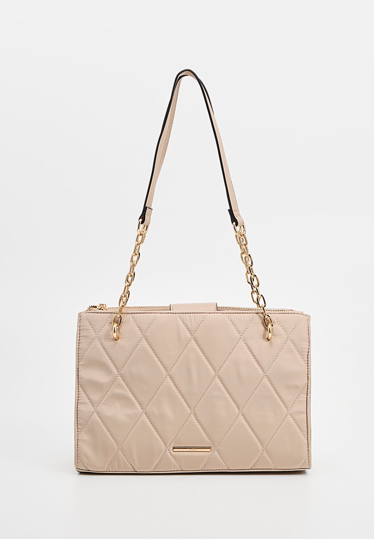 Sac à main matelassé beige clair avec une surface texturée, des anses en chaîne dorée et une fermeture éclair. Présente un petit accent métallique à l'avant.