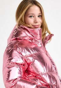 Young woman with blonde hair wearing a shiny metallic pink puffer jacket with high collar, smiling slightly against plain background.