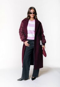 Frau mit schwarzen Sonnenbrillen, einem langen bordeauxfarbenen Mantel, einem gestreiften Pullover in Pink und Weiß, weiten schwarzen Jeans und schwarzen spitzen Schuhen, hält eine kleine bordeauxfarbene Tasche.