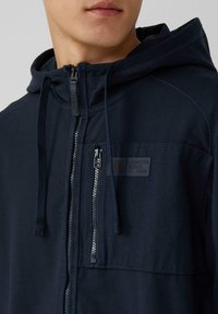 Marineblauwe zip-up hoodie met een trekkoordkap, textuurstof, zijzakken en een logopatch op de borst. De ritshardware is zilverkleurig.