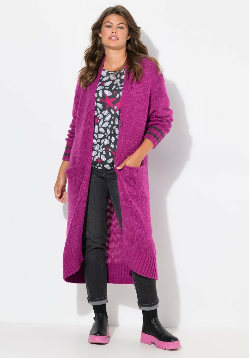 Lange, open voorkardigan in fuchsia breimateriaal met twee voorzakken. Gecombineerd met een patroon top, donkere spijkerbroek en roze platformlaarzen.
