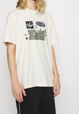 T-shirt en coton beige clair avec un design graphique représentant un canapé, des personnages de cartoon, des plantes en pot et le logo Adidas en violet.