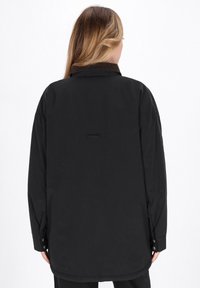 Camisa chaqueta negra de manga larga hecha de una tela suave, con cuello, puños con botones y un pequeño detalle en forma de lazo en la parte trasera.