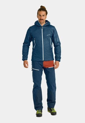 Blue insulated jacket with white zipper, paired with navy pants featuring an orange pocket and reflective accents. Worn with black boots and green laces.