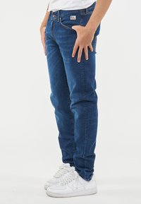 Jeans in denim blu scuro con vestibilità slim, dotati di cinque tasche e un patch con logo cucito sulla cintura. Indossati con sneakers bianche.