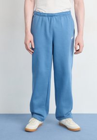 Gaiši zili sporta bikses ar elastīgu jostu, sānu kabatām un mazu baltu logotipu augšējā kājā, apvienotas ar baltām kedām.