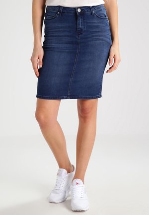 Donna con indosso una gonna di jeans blu scuro e sneaker bianche, in piedi con le gambe incrociate su uno sfondo chiaro uniforme.
