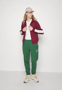 Bordeaux rød lynlåstrøje med hvide ærmer, kombineret med grønne joggingbukser med logo. Beige sneakers og en lys kasket fuldender outfittet.