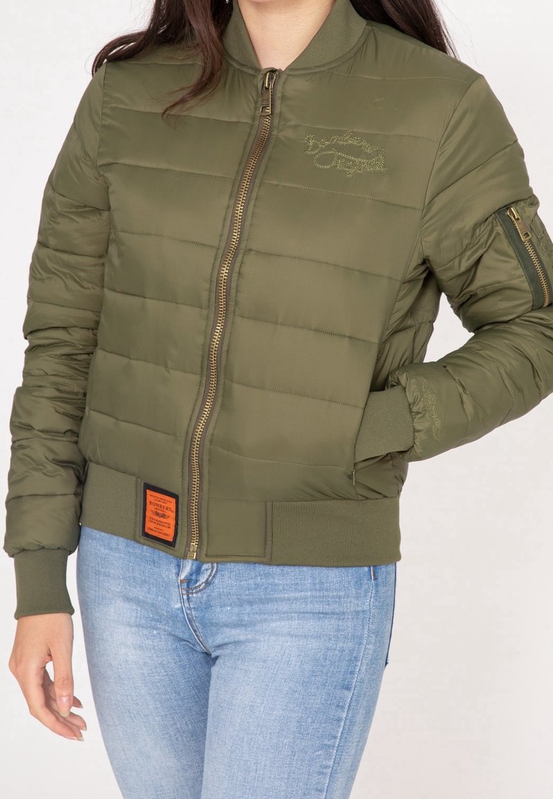 Femme portant une veste bomber matelassée vert olive avec une fermeture éclair devant et un jean bleu clair, main dans la poche côté droit.