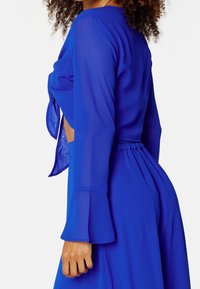 Femme portant un haut transparent, bleu vif, à manches longues noué à l'avant et une jupe assortie taille haute sur un fond blanc.