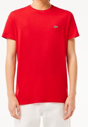 Personne portant un t-shirt rouge uni à manches courtes avec un petit logo crocodile vert sur la poitrine gauche et un pantalon blanc.
