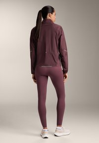Bordeaux lichtgewicht jack met een hoge kraag en reflecterende accenten, gecombineerd met strakke bordeaux leggings en witte sportschoenen.