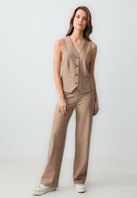 Beige, gestreepte op maat gemaakte vest met een V-hals en vier knopen, gecombineerd met bijpassende losvallende broek. Beide hebben een gladde textuur.