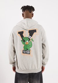 Grauer Kapuzenpullover mit einem großen grünen Drachenmotiv auf der Rückseite, akzentuiert mit einem kräftigen "V" und dem "YRTRN"-Logo in Schwarz und Orange.