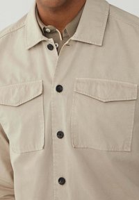 Beige Hemd mit Knopfleiste, zwei aufgesetzten Taschen, schwarzen Knöpfen und einem kragenlosen Ausschnitt. Glatte Textur, leichtes Gewebe und lässiges Design.