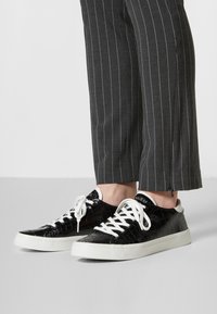 Sneakers neri con texture a coccodrillo, lacci bianchi e suole in gomma, abbinati a pantaloni grigi a righe con una leggera fessura.