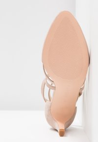 Sandale à talon haut en daim beige avec bout pointu, trois fines brides et semelle texturée, appuyée contre un mur blanc.