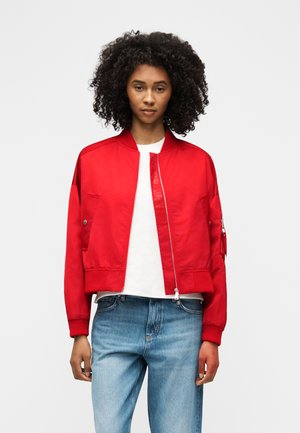 Mujer con cabello rizado que lleva una chaqueta bomber roja brillante sobre una camiseta blanca y jeans azul claro, de pie contra un fondo blanco.