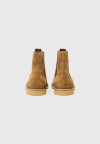 Botas de tobillo de ante marrón con paneles elásticos a los lados, punta redonda y suela beige. La parte trasera cuenta con una tira para facilitar su colocación.