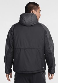 Svart puffjacka med hög krage, synlig sömnad och en Nike-logotyp längst ner till vänster. Slät struktur och något lös passform.