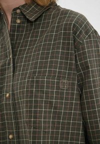 Une chemise à carreaux verte et marron avec un design à boutons, une poche poitrine, et de petits boutons marron clair avec un petit logo sur la poche.