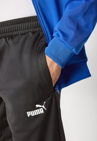 Blå lynlåsjakke og sorte bukser, begge med en glat overflade. De sorte bukser har et hvidt Puma-logo på benet.