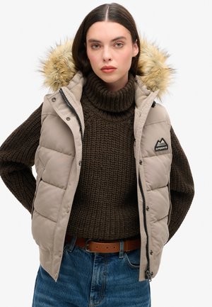 Gilet matelassé beige avec capuche en fourrure, fermeture éclair noire et logo de montagne. Porté par-dessus un pull en tricot marron foncé et un jean bleu.