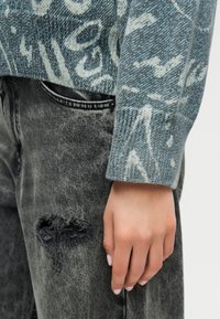 Χέρι που ακουμπάει στο πλάι, φορώντας μπλε πουλόβερ με σχέδιο πλέξης και μαύρο τζιν με φθορές, με ορατή τσέπη.