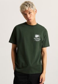 Camiseta de algodón verde oscuro con un gráfico blanco que presenta el texto "VANS" y "OFF THE WALL". Mangas cortas, cuello redondo, ajuste casual.