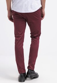 Muž v slim-fit burgundských kalhotách, černých botách a bílém tričku, stojící s uvolněnýma rukama podél těla na čistém bílém pozadí.