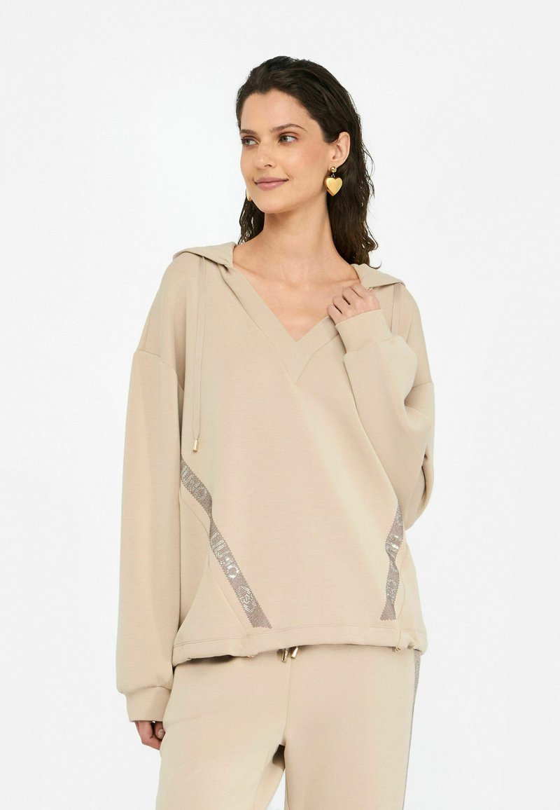 Haut à capuche beige oversize avec un col en V, capuche à cordon de serrage, et accents de sequins sur les côtés. Texture douce et coupe décontractée.