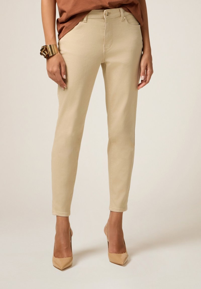 Pantalones beiges de corte slim con una textura suave, diseño de cinco bolsillos y cierre frontal con botones, combinados con tacones beiges de punta afilada y una blusa marrón.