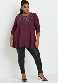 Top bordeaux a maniche lunghe con scollatura a V e dettagli di perline sul collo, abbinato a leggings neri aderenti e lucidi e tacchi neri con cinturini.