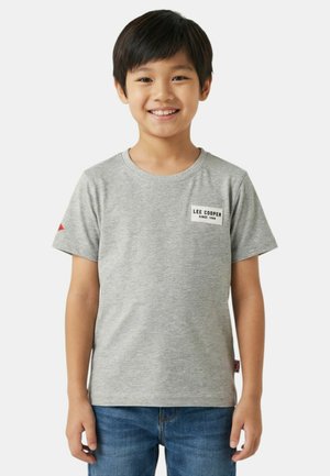 Garçon souriant portant un t-shirt Lee Cooper gris clair et un jean bleu, posé devant un fond blanc uni, les bras détendus.