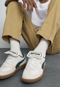 Baskets blanches en cuir avec des accents verts, des détails texturés sur les côtés, une languette rembourrée et une semelle en caoutchouc. Portées avec un pantalon beige et des chaussettes blanches.