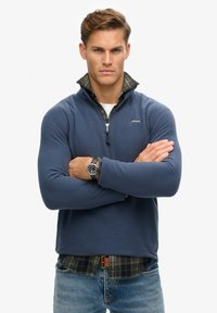 Un pulóver de forro polar azul con un cuello alto, cierre de cremallera y una camisa de cuadros negra y verde debajo. Combinado con unos vaqueros azul claro y un reloj de pulsera.