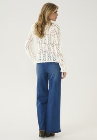 Top bianco in pizzo a maniche lunghe con intricate aperture e motivi, abbinato a jeans in denim blu a gamba larga. Design minimalista, tonalità neutre.