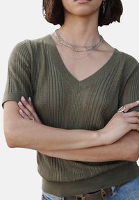 Haut tricot à manches courtes de couleur vert olive avec un col en V, présentant des motifs à côtes verticales et un ourlet ajusté. S'accompagne de colliers en argent superposés.