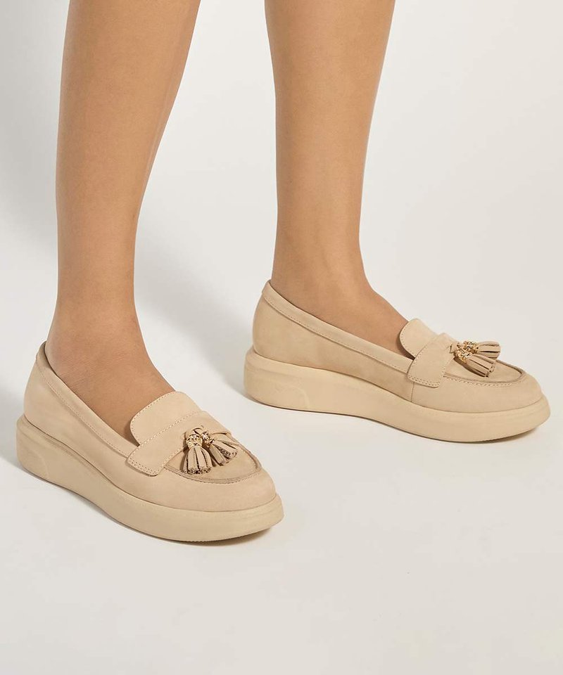 Femme portant des mocassins à plateforme beige sans lacets avec des détails pompons sur un fond neutre.