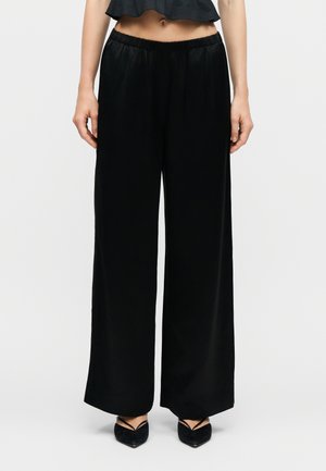 Kvinde iført sorte bukser med vide ben og elastisk talje, kombineret med sorte spidse hæle og en kort sort top.