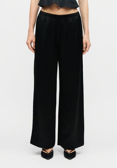 Femme portant un pantalon noir à jambes larges avec une taille élastique, associée à des talons noirs pointus et un haut noir court.