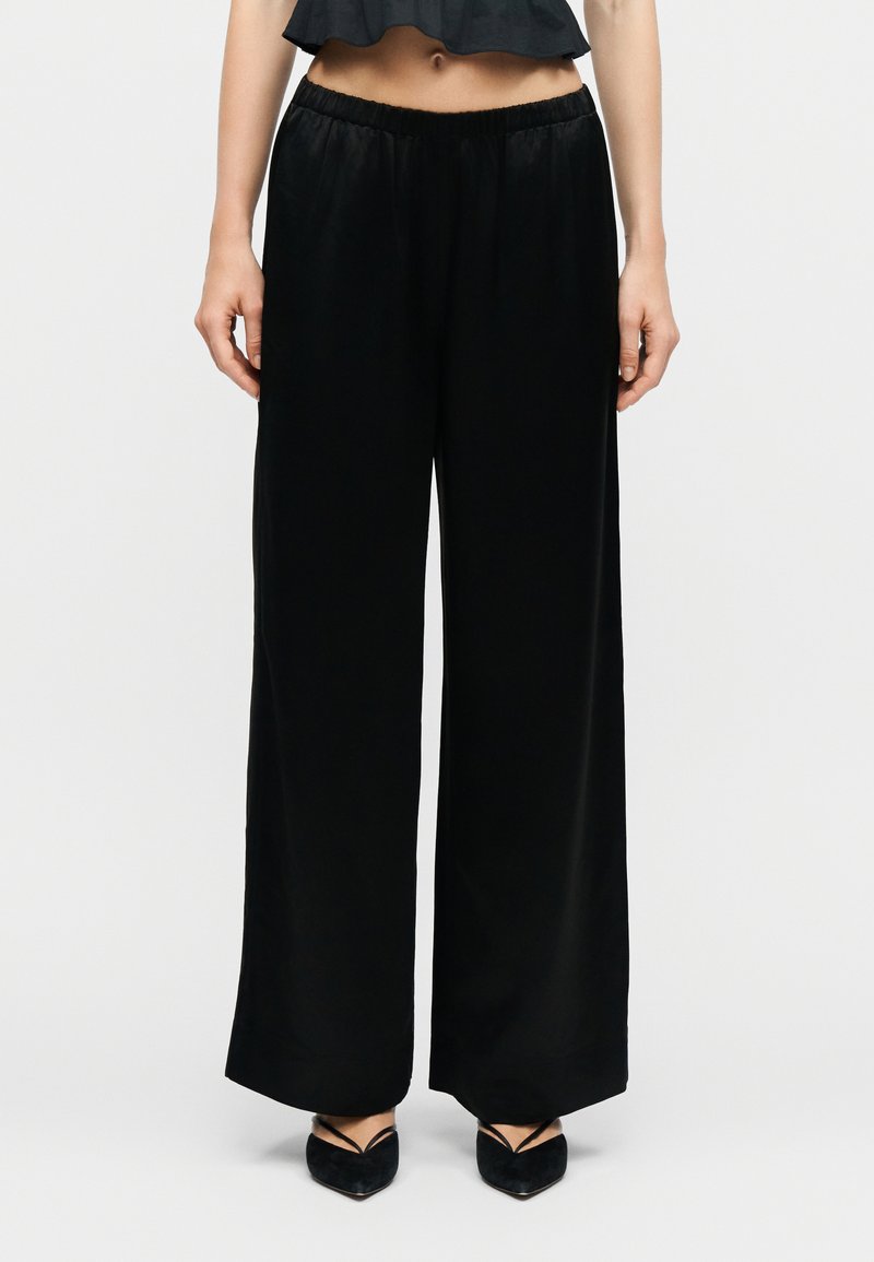 Γυναίκα που φοράει μαύρο παντελόνι με φαρδιά μπατζάκια και ελαστική μέση, συνδυασμένο με μαύρες μυτερές γόβες και μαύρο cropped μπλουζάκι.